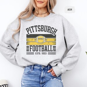 May include: Light gray sweatshirt with the text "PITTSBURGH FOOTBALL ESTD. 1933" in black and gold. The design includes a football graphic. The sweatshirt is a comfortable, casual style.