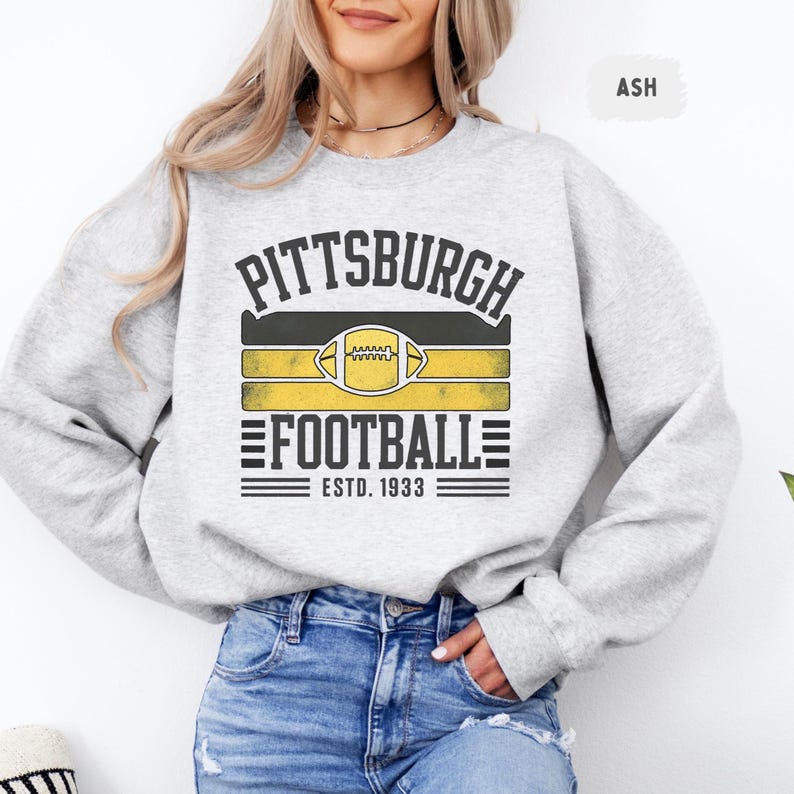 May include: Light gray sweatshirt with the text "PITTSBURGH FOOTBALL ESTD. 1933" in black and gold. The design includes a football graphic. The sweatshirt is a comfortable, casual style.