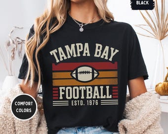 Puede incluir: Camiseta negra con "TAMPA BAY FOOTBALL" en letras blancas sobre un gráfico de fútbol americano. El gráfico está sobre un fondo de rayas rojas, naranjas y doradas. El texto "ESTD. 1976" está debajo.