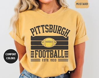 May include: Mustard-coloured t-shirt featuring a Pittsburgh Football graphic. The design includes the text "PITTSBURGH FOOTBALL ESTD. 1933" and a football graphic. The shirt is a comfort colour.