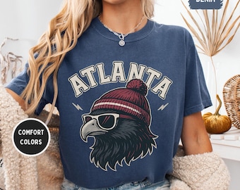 May include: Denim blue t-shirt with the word "ATLANTA" in white arched text. The shirt features a graphic of a bird wearing sunglasses and a maroon beanie. The words "COMFORT COLORS" are in a white circle.