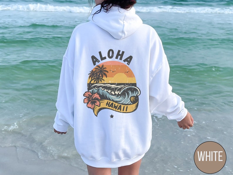 Puede incluir: Sudadera con capucha blanca con un estampado gr&aacute;fico de una puesta de sol sobre una ola con palmeras y el texto "ALOHA HAWAII".