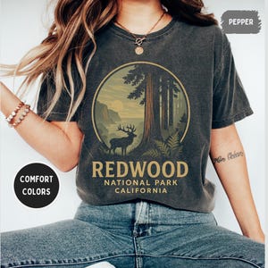 Pode incluir: Camiseta cor pimenta com um gráfico circular de uma floresta de sequoias com um veado. O texto "REDWOOD NATIONAL PARK CALIFORNIA" está impresso abaixo. A camiseta é de cor confortável.
