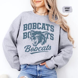 Puede incluir: Sudadera gris deportiva con la palabra "BOBCATS" en una fuente universitaria y un gr&aacute;fico verde azulado de un lince pardo gru&ntilde;endo. La sudadera es de cuello redondo y est&aacute; hecha de un material suave.