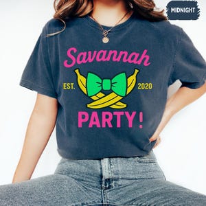 May include: A navy blue t-shirt with the text "Savannah PARTY!" in pink. The design includes a green bow and yellow bananas. The text "EST. 2020" is also present. The shirt is a casual style.
