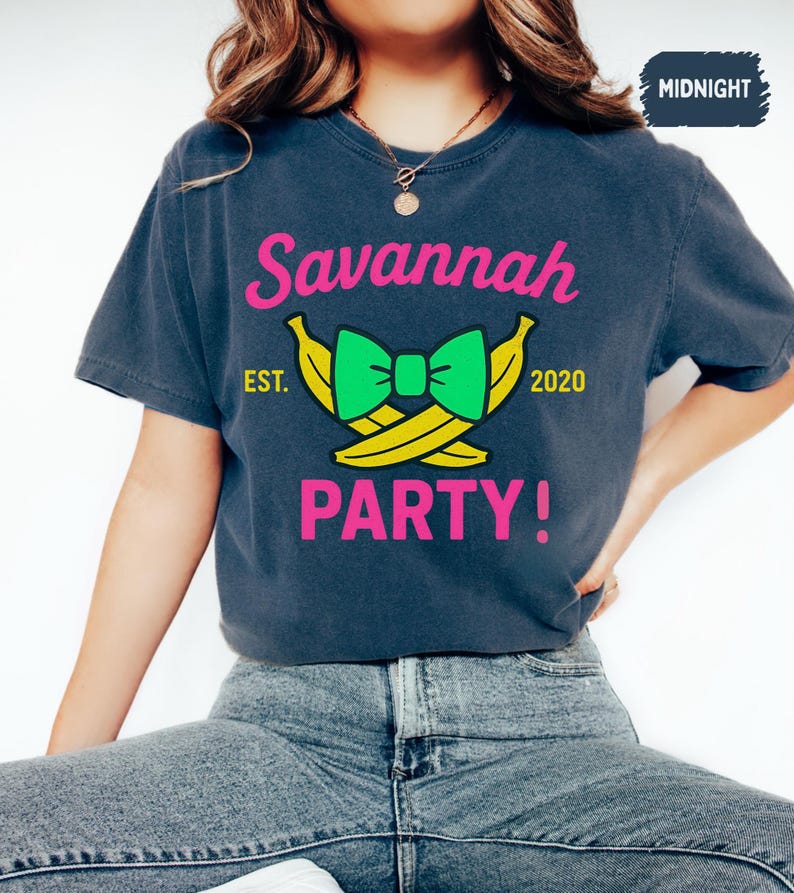 May include: A navy blue t-shirt with the text "Savannah PARTY!" in pink. The design includes a green bow and yellow bananas. The text "EST. 2020" is also present. The shirt is a casual style.