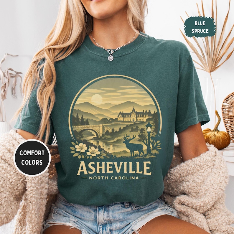 May include: A teal-colored t-shirt featuring a circular graphic of Asheville, North Carolina. The design includes a bridge, mountains, a deer, and the text "Asheville - North Carolina". The shirt also has the text "Comfort Colors" and "Blue Spruce".