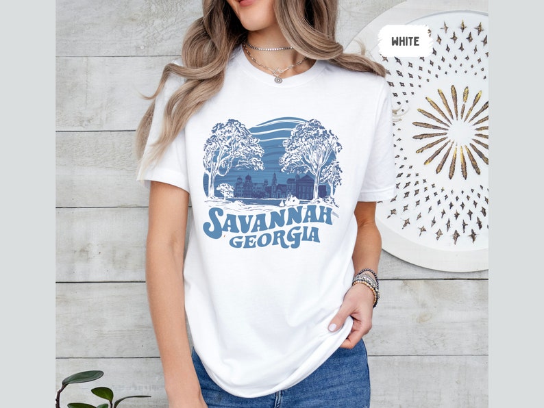 Puede incluir: Camiseta blanca con un dise&ntilde;o gr&aacute;fico azul de una ciudad con &aacute;rboles y el texto "SAVANNAH GEORGIA".