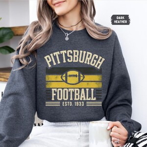 May include: Dark heather gray sweatshirt with "Pittsburgh Football" text and a football graphic in yellow and black. The text "ESTD. 1933" is below the graphic. The sweatshirt is a crew neck style.