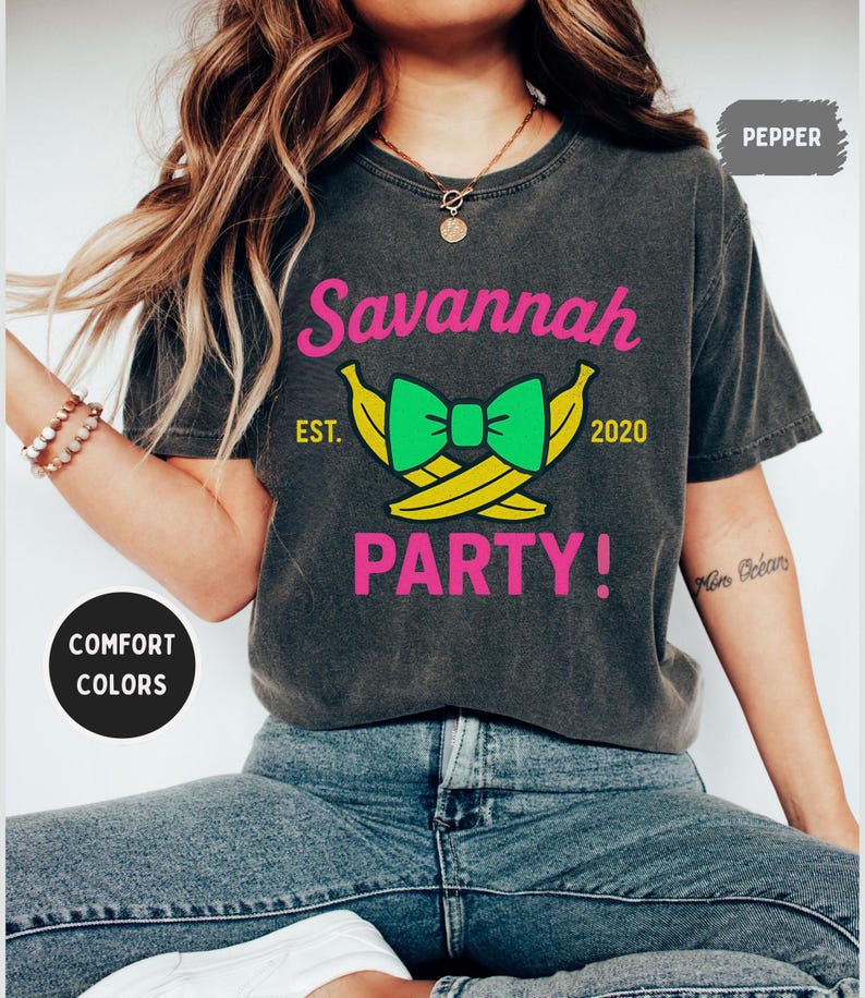 May include: Dark gray t-shirt with the words "Savannah PARTY!" in pink, and a green bow tie over two yellow bananas. The text "EST. 2020" is also included. The shirt is a comfort color.