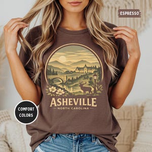 May include: Espresso-colored t-shirt featuring a circular graphic of a mountain landscape with a bridge, deer, and the text "Asheville North Carolina." The shirt has a relaxed fit and the words "Comfort Colors" are printed on the shirt.