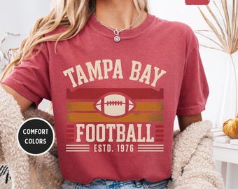 Puede incluir: Camiseta granate con las palabras "TAMPA BAY FOOTBALL" y "ESTD. 1976" impresas en un estilo vintage. El diseño incluye un gráfico de fútbol americano y rayas horizontales en tonos rojos y dorados. La camiseta tiene cuello redondo.