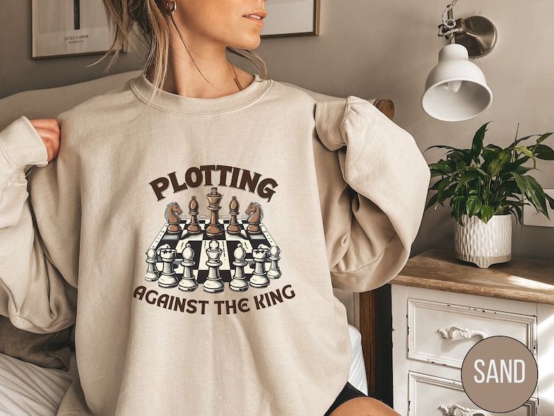 May include: A beige sweatshirt with a brown graphic of a chessboard and the text "Plotting Against The King".
