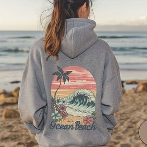 Op de afbeelding: Grijze hoodie met een strandafbeelding op de rug. Het ontwerp toont een palmboom, een golf en de woorden "Ocean Beach". De hoodie is neutraal van kleur en geschikt voor dagelijks gebruik. De tekst "Sport Gray" is ook zichtbaar.