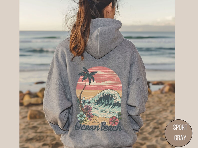 Op de afbeelding: Grijze hoodie met een strandafbeelding op de rug. Het ontwerp toont een palmboom, een golf en de woorden "Ocean Beach". De hoodie is neutraal van kleur en geschikt voor dagelijks gebruik. De tekst "Sport Gray" is ook zichtbaar.