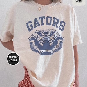 May include: An ivory short-sleeve t-shirt featuring a distressed blue graphic of an alligator head centred below the text GATORS in a collegiate font. A circular black badge on the left reads COMFORT COLORS and a small tag in the top right reads IVORY.