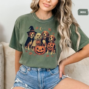 Op de afbeelding: Mosgroen t-shirt met een Halloween-ontwerp. De afbeelding toont vijf honden in kostuums, een uitgesneden pompoen, een halve maan en herfstbladeren. Het woord "MOSS" staat rechtsboven.