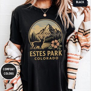 May include: Black t-shirt with a circular graphic featuring a mountain range, a deer, and a flower. The text "ESTES PARK COLORADO" is below the graphic. The shirt also has the text "COMFORT COLORS" on a circular sticker.