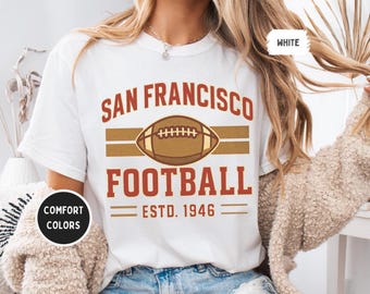 May include: White t-shirt featuring a San Francisco Football design in brown and gold. The design includes a football graphic and the text "ESTD. 1946". The shirt is a comfort colors style.
