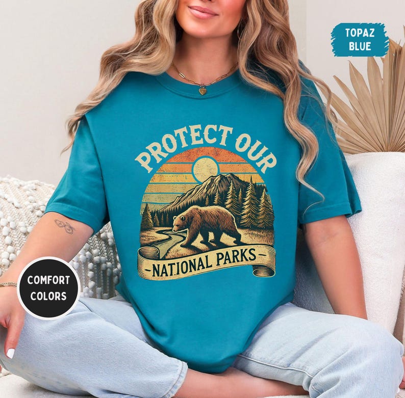 May include: A turquoise blue t-shirt with a vintage-style graphic of a bear in a forest scene. The graphic says "Protect Our National Parks".