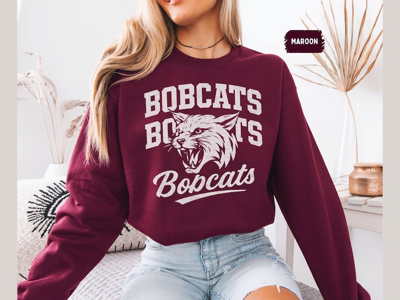 Puede incluir: Sudadera granate con la palabra "BOBCATS" en letras blancas, con un gr&aacute;fico de cabeza de gato mont&eacute;s gru&ntilde;endo. La palabra "Bobcats" se repite debajo del gr&aacute;fico. La palabra "MAROON" est&aacute; en un peque&ntilde;o cuadro en la esquina superior derecha.