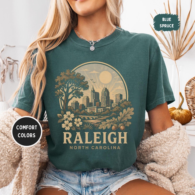 May include: A teal-colored t-shirt featuring a circular graphic of the Raleigh, North Carolina skyline, a tree, and the text "RALEIGH NORTH CAROLINA." The shirt has a crew neck and short sleeves. The words "Comfort Colors" are printed on a black circle.