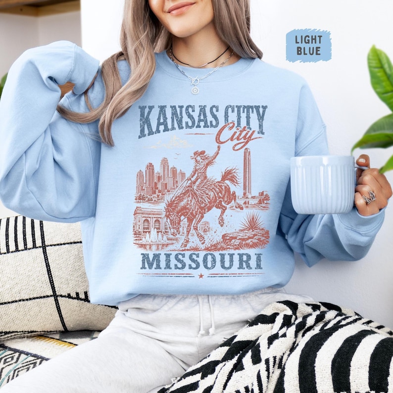 May include: Light blue sweatshirt featuring a vintage-style graphic of Kansas City, Missouri. The design includes a cowboy riding a bucking bronco, city skyline, and the text "Kansas City Missouri". The sweatshirt has a crew neck and long sleeves.