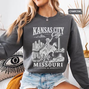 May include: Charcoal gray sweatshirt featuring a white graphic of a cowboy riding a bucking bronco, with the text "KANSAS CITY City MISSOURI". The sweatshirt has a crew neck and long sleeves. The word "CHARCOAL" is printed in the upper right corner.