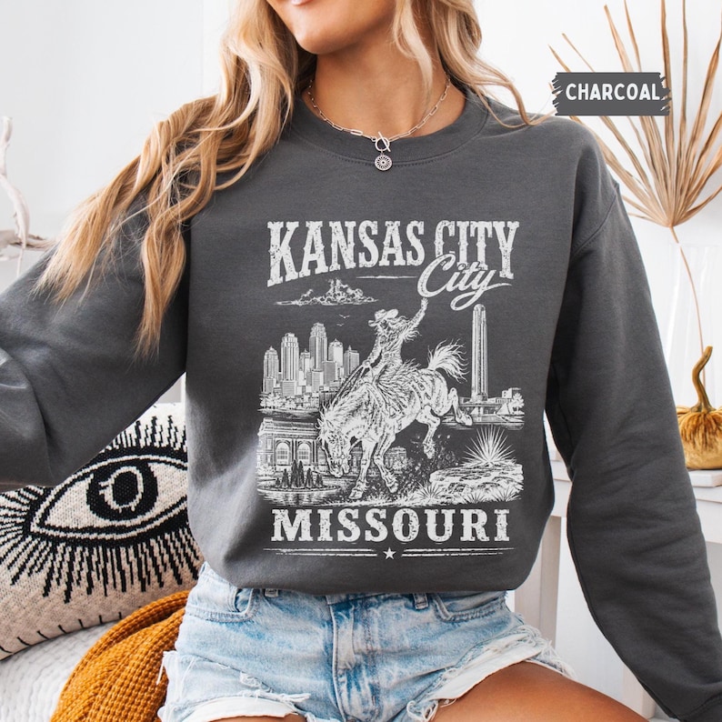 May include: Charcoal gray sweatshirt featuring a white graphic of a cowboy riding a bucking bronco, with the text "KANSAS CITY City MISSOURI". The sweatshirt has a crew neck and long sleeves. The word "CHARCOAL" is printed in the upper right corner.