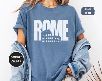 May include: Blue jean-colored t-shirt with the word "ROME" in large white letters and a white graphic of the Colosseum. The shirt is a Comfort Colors brand. The text "BLUE JEAN" is in a blue rectangle.