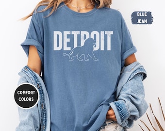 May include: A blue jean colored t-shirt with the word "DETROIT" in white block letters and a white lion graphic. The shirt has rolled-up sleeves and is part of the "Comfort Colors" collection.