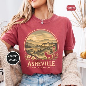 May include: A maroon t-shirt with a circular graphic featuring a scenic landscape of Asheville, North Carolina. The design includes a bridge, mountains, a deer, and the text "Asheville - North Carolina". The shirt also has the text "Comfort Colors" and "Crimson".