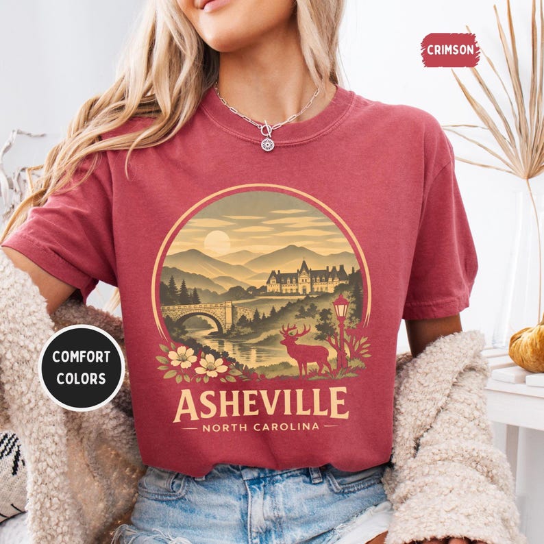 May include: A maroon t-shirt with a circular graphic featuring a scenic landscape of Asheville, North Carolina. The design includes a bridge, mountains, a deer, and the text "Asheville - North Carolina". The shirt also has the text "Comfort Colors" and "Crimson".