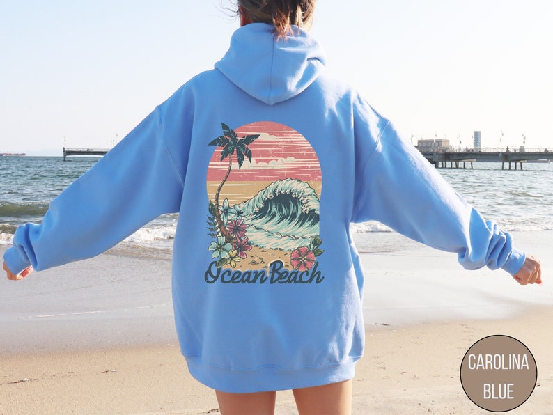 Op de afbeelding: Lichtblauwe hoodie met een retro graphic op de rug. Het ontwerp toont een palmboom, een oceaangolf en de woorden "Ocean Beach". De graphic bevat roze, blauwe en groene bloemaccenten. De hoodie is lichtblauw.