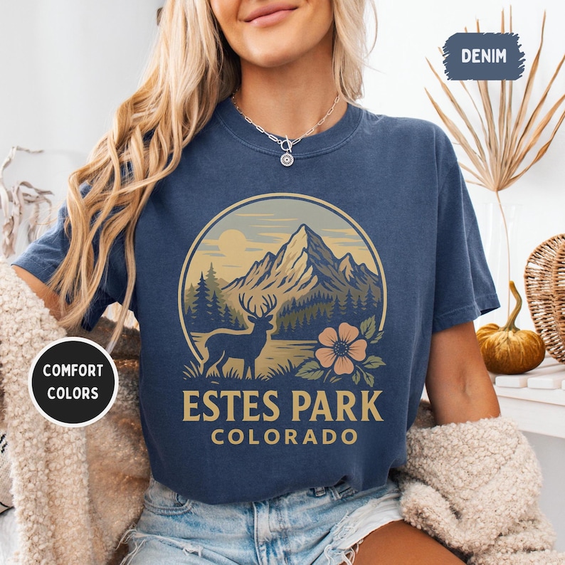May include: A navy blue t-shirt featuring a circular graphic of a mountain range, a deer, and a flower. The text "Estes Park Colorado" is printed below the graphic. The shirt has short sleeves and a crew neck.