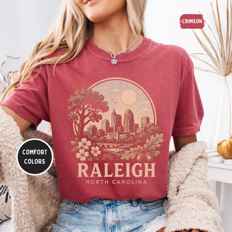 May include: Crimson-colored t-shirt featuring a circular graphic of the Raleigh, North Carolina skyline, with a tree, flowers, and the sun. The word "RALEIGH" is printed below the graphic, with "NORTH CAROLINA" underneath. The shirt has the words "COMFORT COLORS" printed on it.