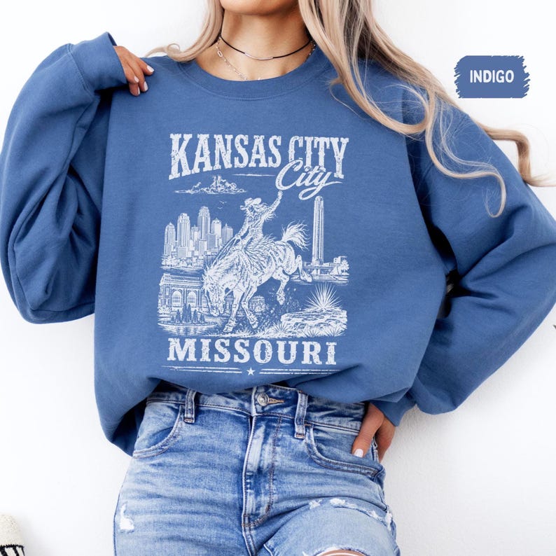 May include: Indigo blue sweatshirt featuring a white graphic of Kansas City, Missouri. The design includes the city skyline, a bucking bronco rider, and the text "KANSAS CITY City MISSOURI". The sweatshirt has long sleeves and a crew neck.