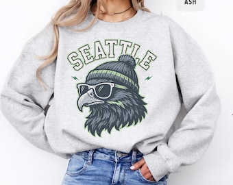 Könnte beinhalten: Hellgraues Sweatshirt mit einem Aufdruck eines Adlers mit Sonnenbrille und Mütze. Das Wort "SEATTLE" ist in Grün und Weiß über dem Adler aufgedruckt. Das Sweatshirt ist aus weichem Material.