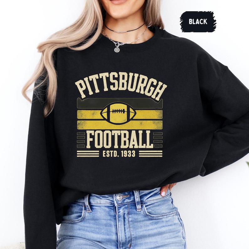 May include: Black sweatshirt with a Pittsburgh Football design. The design features the words "Pittsburgh Football" above and below a football graphic, with "ESTD. 1933" below. The design is in gold and black.