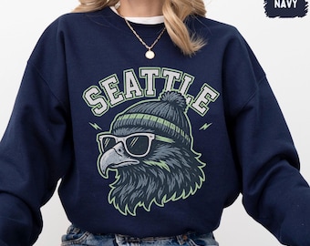 Könnte beinhalten: Marineblaues Sweatshirt mit einer Grafik eines Adlers mit Sonnenbrille und Mütze. Das Wort "SEATTLE" ist in grüner und weißer Schrift über dem Adler angeordnet. Das Sweatshirt hat einen Rundhalsausschnitt und lange Ärmel.