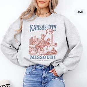 May include: Ash-colored sweatshirt featuring a distressed graphic of a rodeo rider on a bucking bronco, with the words "Kansas City Missouri" in blue and red. The design includes city skyline and architectural elements.