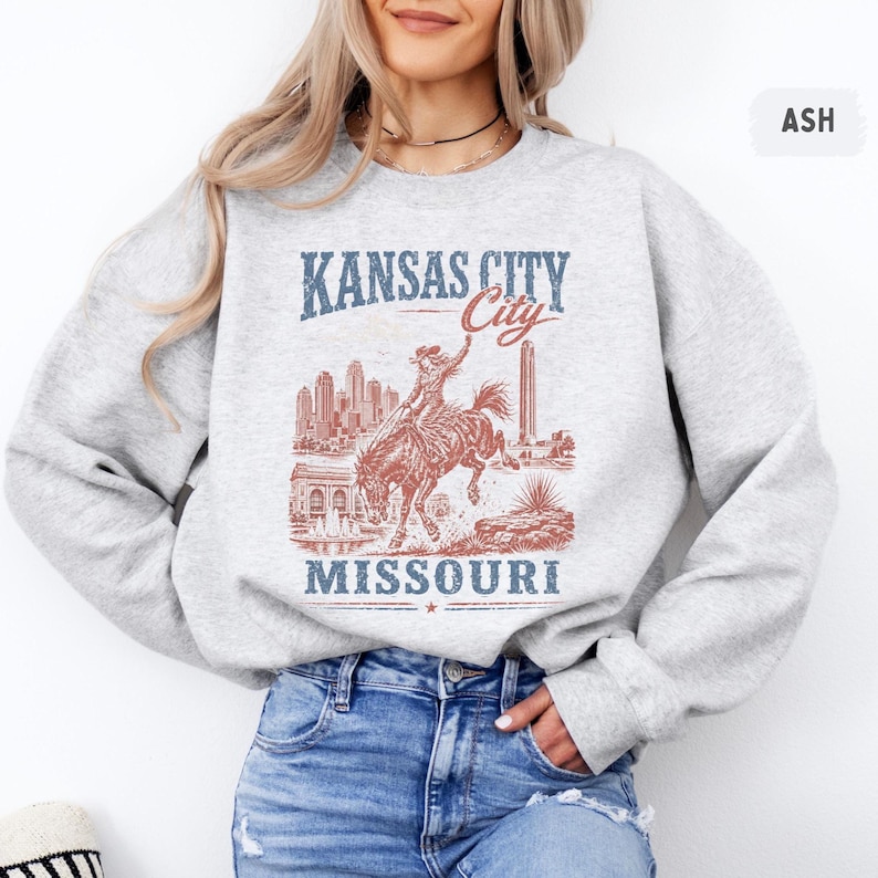May include: Ash-colored sweatshirt featuring a distressed graphic of a rodeo rider on a bucking bronco, with the words "Kansas City Missouri" in blue and red. The design includes city skyline and architectural elements.