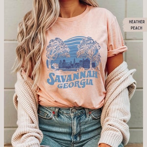 Puede incluir: Una camiseta de color melocot&oacute;n jaspeado con un gr&aacute;fico azul de una ciudad con &aacute;rboles y el texto "Savannah Georgia".