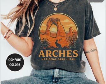 May include: Dark grey t-shirt featuring a circular graphic of Arches National Park in Utah. The design includes an arch, a rabbit, and the text "ARCHES NATIONAL PARK - UTAH" in orange. The shirt is a comfort color.