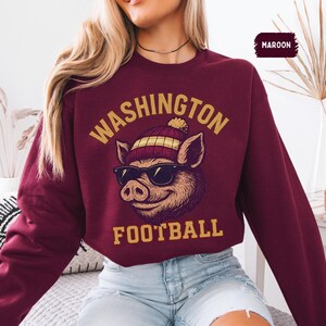 May include: Maroon sweatshirt with the text "WASHINGTON FOOTBALL" in gold lettering. The graphic features a pig wearing sunglasses and a knit cap. The sweatshirt is a solid maroon color.
