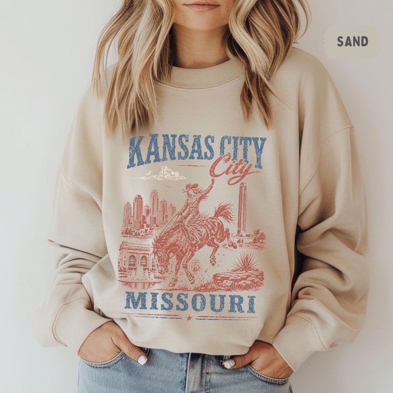 May include: Sand-colored sweatshirt featuring a distressed graphic of Kansas City, Missouri. The design includes a cowboy riding a bucking bronco, city skyline, and the text "Kansas City" and "Missouri" in blue and red.