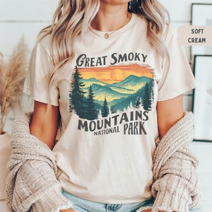 May include: A cream colored t-shirt with a vintage style graphic of a mountain range with a sunset and the text "Great Smoky Mountains National Park".