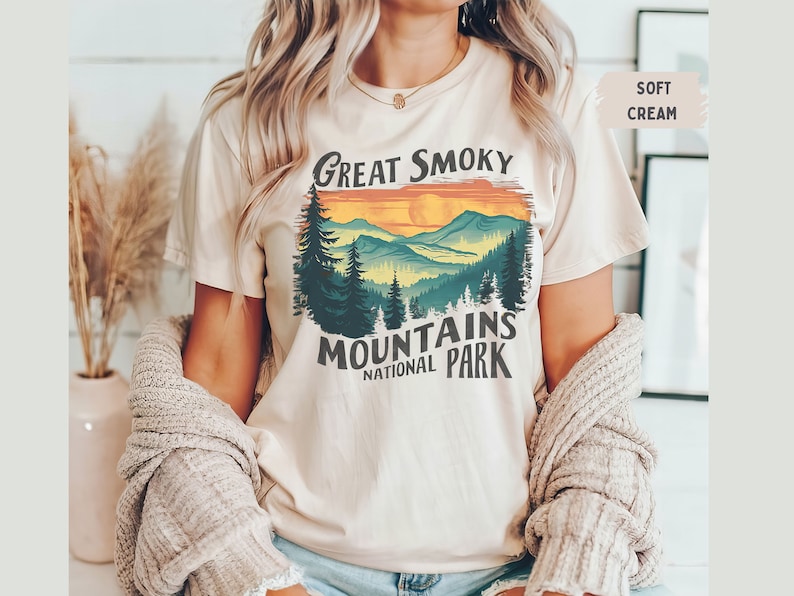 May include: A cream colored t-shirt with a vintage style graphic of a mountain range with a sunset and the text "Great Smoky Mountains National Park".