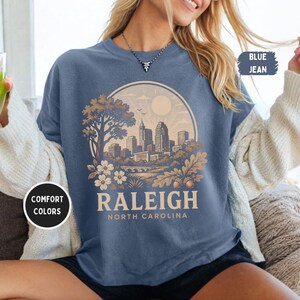 May include: Blue jean-colored t-shirt with a graphic of the Raleigh, North Carolina skyline inside a circular design. The design includes a tree, flowers, and the text "RALEIGH NORTH CAROLINA". The shirt also has the text "COMFORT COLORS".