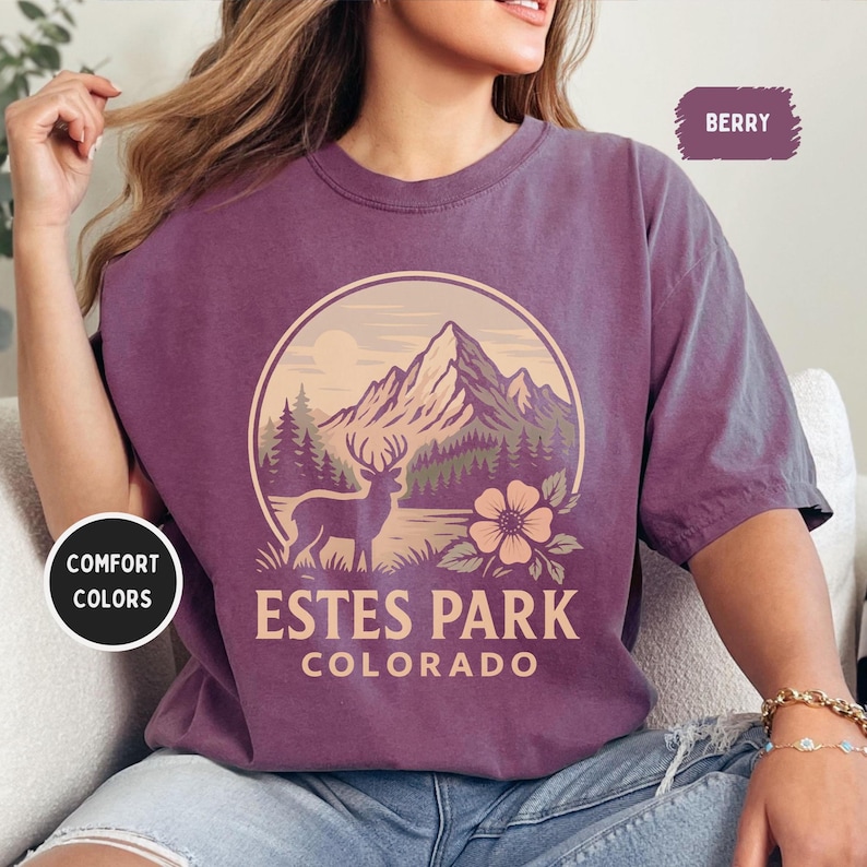 May include: Berry-colored t-shirt with a circular graphic featuring a mountain range, deer, and flower, with the text "Estes Park Colorado." The shirt also has the text "Comfort Colors."